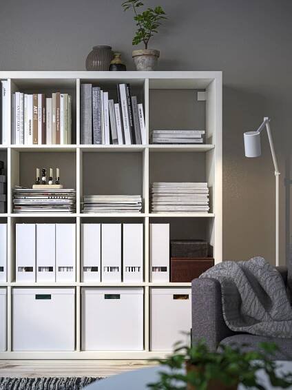 Ein weißes KALLAX Regal 4x4 mit Zeitschriftensammlern, Büchern und weißen Papierboxen steht an einer Wohnzimmerwand. 