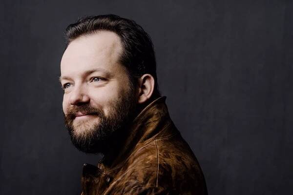 Portrait von Andris Nelsons. Mann mit dunklen Haaren und Vollbart steht vor schwarzem Hintergrund und richtet den Blick nach rechts