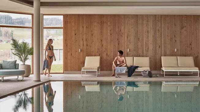 Zwei Personen in Wellness-Umgebung mit Holzwand, Liegen und Pool mit Reflexionen.