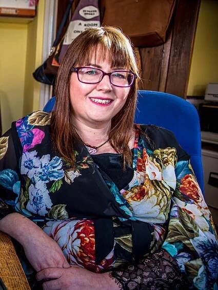 Medium shot of Rebecca Roberts. Rebecca is sitting at her desk at home in a yellow room. She's wearing a black dress with a colourful flower print, rectangle dark red glasses and has medium-length brown hair.