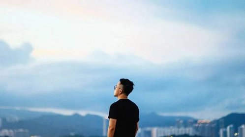 A male standing agains the backdrop of sky, mountains, and skyscrapers.