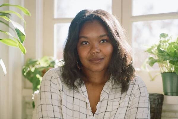 A portrait of Courtney Faye Taylor sitting in a sunlit room, surrounded by plants.
