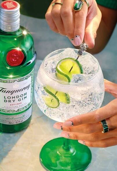 A Gin & Tonic cocktail made with Tanqueray London Dry Gin garnished with two lime slices. A bottle of Tanqueray London Dry gin sits in the background of the image.