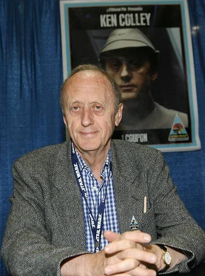 A gaunt man wearing a sport coat and a blue checked shirt looks forward with his hands clasped in front of him. Behind him is a poster of his character, Adm. Firmus Piett, a grim-faced man in a military uniform.