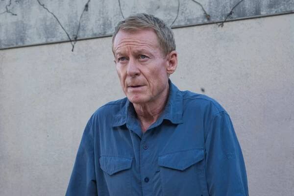 A film still of Richard Roxburgh, a middle-aged blond man, in a blue button-down, with his hands on a car bonnet.