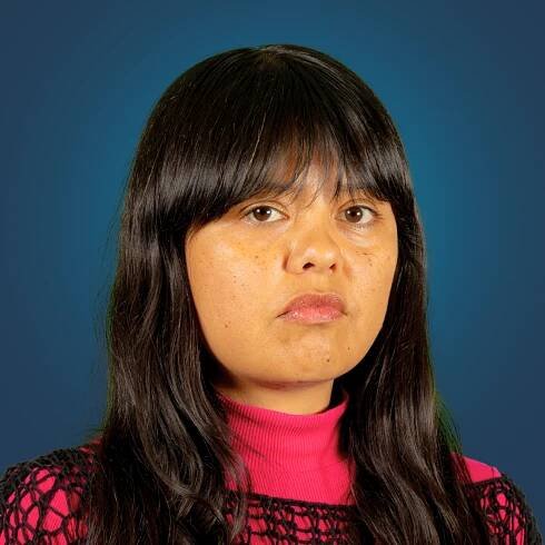 A woman with long black hair is posing seriously in front of a blue background. She wears a pink turtleneck sweater and a black netted top, with a neutral expression.