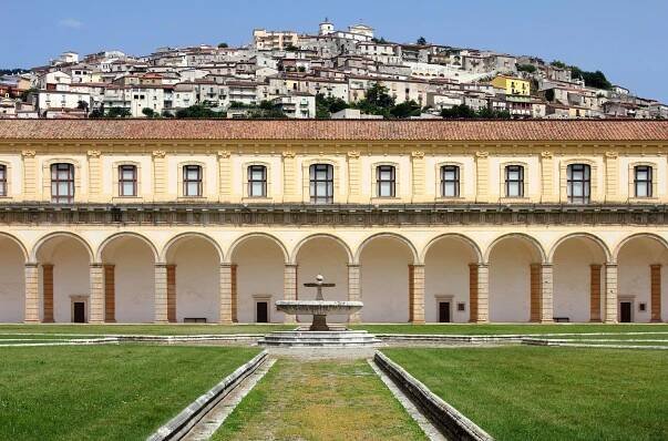 Historic courtyard with arches and a central fountain, overlooking a hillside town in Italy. The landscape features a blend of architectural styles and lush greenery, showcasing the region's cultural heritage.