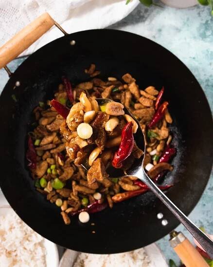 A spoon of vegan kung pao chicken being lifted from a pan showing the chilli and peanuts in it.