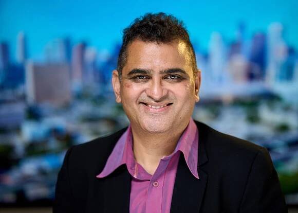 Professional headshot against city skyline backdrop featuring person in black suit and purple shirt.
