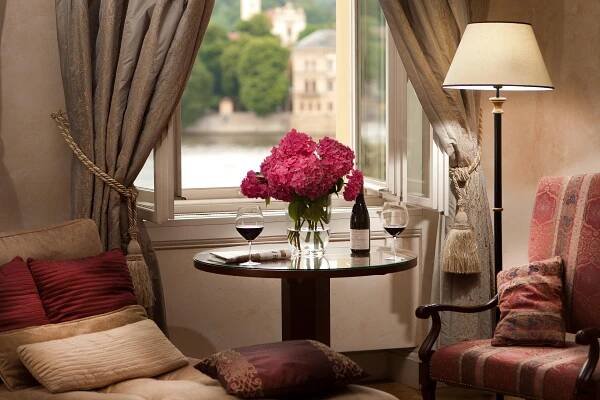 Hotel The Mozart Praha Pokoj fotografie The photo shows a cozy and inviting corner of a room, likely a hotel or a well-appointed home. The focal point is a small, round glass table positioned near a large window. On the table, there's a vase filled with vibrant pink hydrangeas, a bottle of