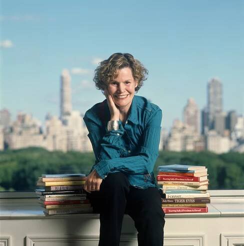 Judy Blume sits on a window ledge, books on either side of her, the New York City skyline behind her. 