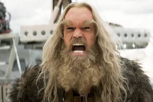 A bearded man with long hair and fur clothing, appearing angry, in a dramatic scene from a movie or TV show