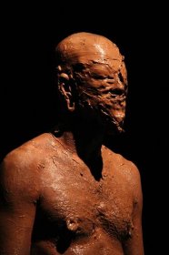 The artist's torso and head covered in wet brown clay. His eyes and mouth are obscured by the clay.