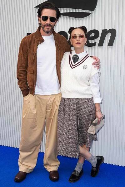 Two people standing together at an event with one person wearing a brown jacket and light pants and the other in a white sweater and plaid skirt