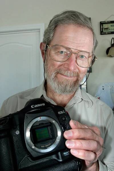 George Smith, an older man with grayish hair, a graying beard and glasses, holds up a Canon camera.