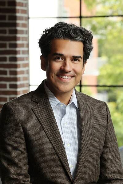 Professional headshot of a man in brown textured suit jacket against brick wall.