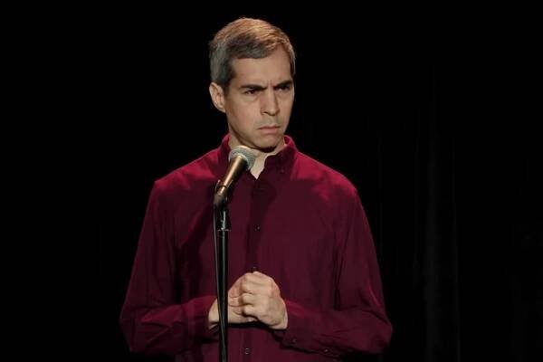 In a shot from a comedy special, a man in a red shirt stands onstage in front of a microphone, wearing a stern look. 