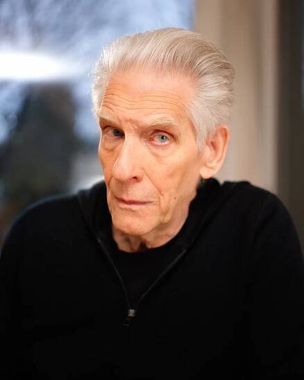 An older man with neatly combed white hair and blue eyes looks thoughtfully at the camera. He is wearing a black zip-up hoodie, and the background is softly blurred with hints of light.