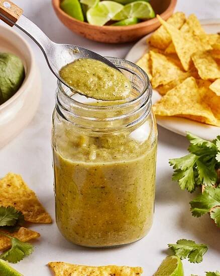 A spoon holding green salsa above a glass jar, surrounded by tortilla chips, lime wedges, cilantro, and an avocado on a white surface.