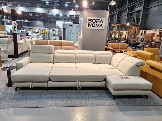 V showroomu je pod nápisem "SOFA NOVA" vystavena velká moderní bílá sekční pohovka s nastavitelnými opěrkami hlavy. Pohovka je vybavena lehátkem a je obklopena dalšími různými pohovkami a kusy nábytku.