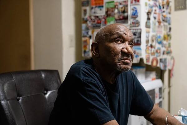 Raymond Johnson sits in a black desk chair in his home. Behind him, out of focus, a white appliance is adorned with colorful magnets and pictures.