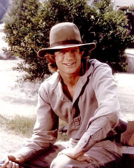 Dean Butler wearing a wide-brimmed hat and a casual button-up shirt, sits outdoors, smiling at the camera