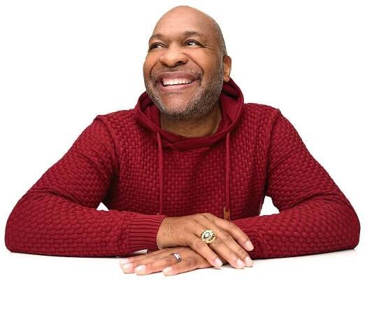 A man in a red sweater, sitting with his arms resting on a white surface, smiling and looking slightly upward.