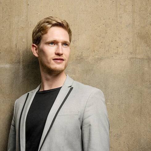 Junger Mann mit blondem Haar und leichtem Bart, in einem hellgrauen Sakko mit schwarzem Shirt, vor einer strukturierten Betonwand. Sein Blick geht leicht zur Seite.