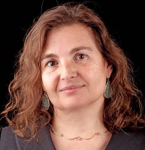 a woman with short, curly reddish brown hair with gold and green teardrop earrings