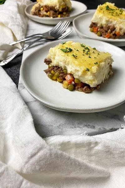 Vertical image of three plates with a hefty serving each of shepherd's pie next to white towels and metal forks.