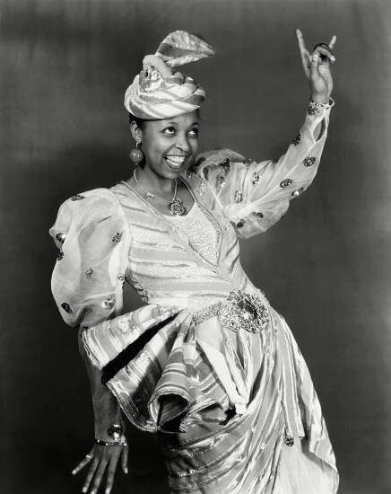 In a black-and-white photo, the actress and blues singer Ethel Waters is grinning and posing with her left arm pointed to the sky, looking upwards in a beaded dress and matching hat. 