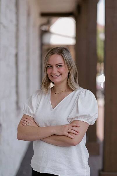 Blonde woman with arms crossed, smiling in front of a brick and column backdrop.