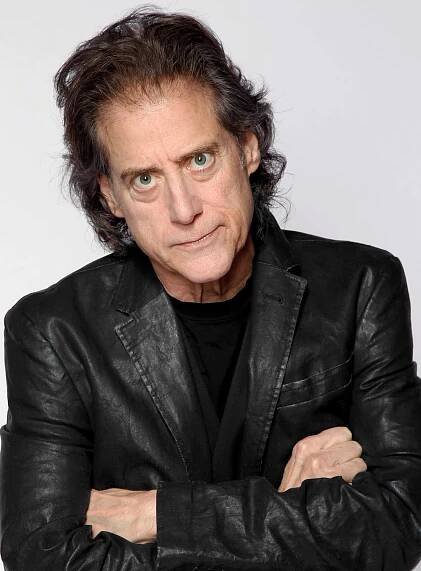 Richard Lewis, an intense-looking dark-haired man wearing a black leather jacket over a black T-shirt with his arms crossed.