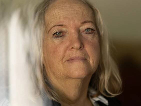 A close-up of a woman's face looking into the camera with a neutral expression. She has long grey hair and blue eyes. 