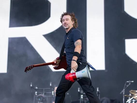 Tim McIlrath holding a megaphone in one hand and his guitar in the other. He is walking across a stage with a smile on his face.