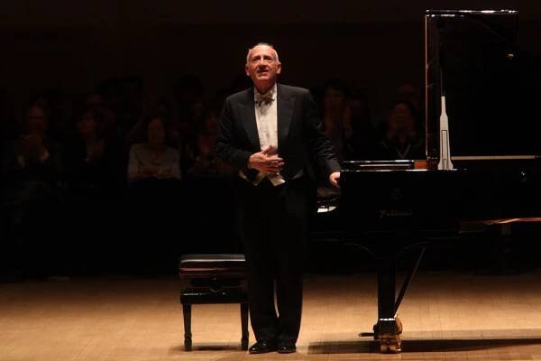 Maurizio Pollini, a white-haired older man in a tuxedo, stands at a piano on a concert stage and acknowledge the audience’s applause.