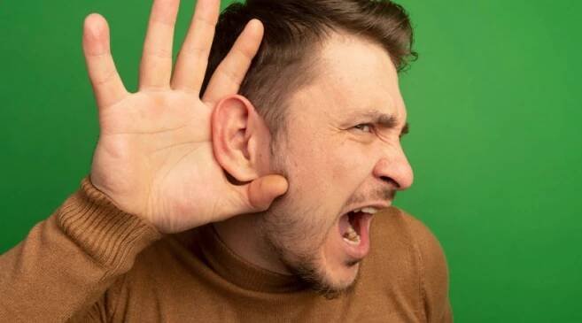 Close up view curious young blonde handsome man keeping hand ear looking straight doing i can t hear you gesture