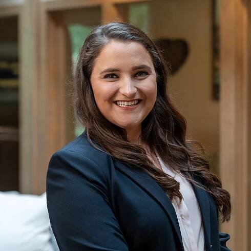Headshot of a smiling young woman with long, brunette hair dressed in a blazer in front of a blurred background