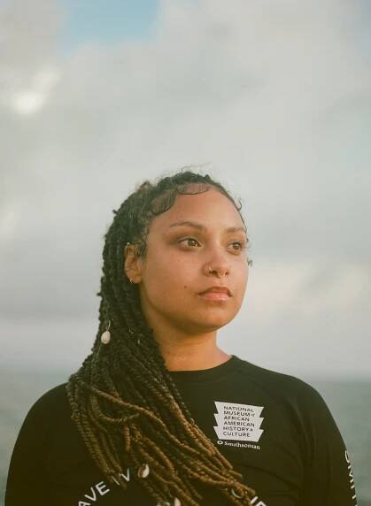 Headshot of a woman with long braids wearing a black shirt with a background of the sky and clouds.