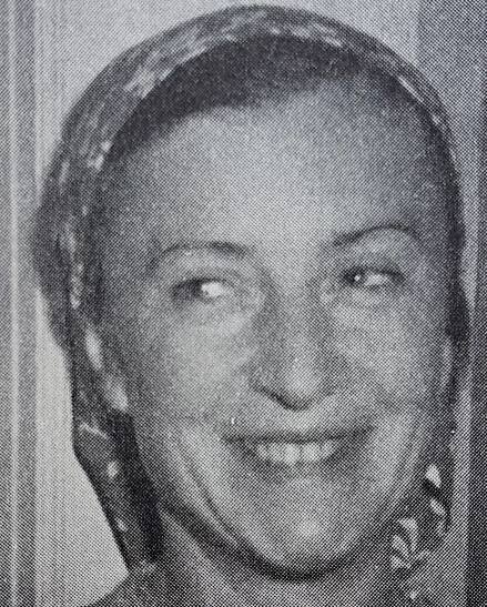 Close-up in black and white of a smiling woman. She has tied a scarf around her hair.