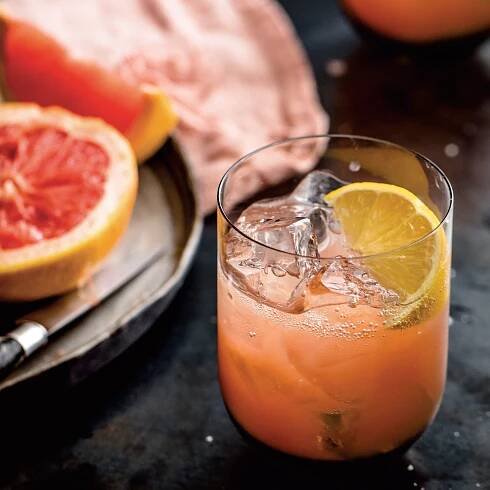 A lightpink cocktail with a lemon slice in a small glass next to fresh grapefruit.