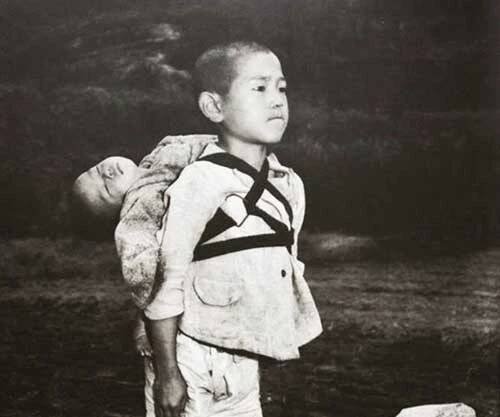 A Japanese boy standing at attention after having brought his dead younger brother to a cremation pyre, 1945