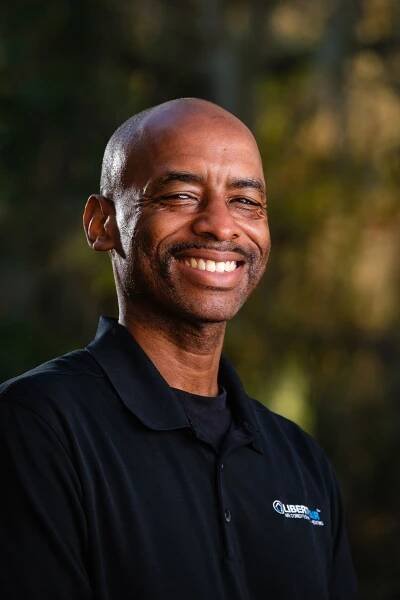 Corporate headshot of a smiling man with a shaved head in a black polo shirt outdoors in Gainesville, Fl.