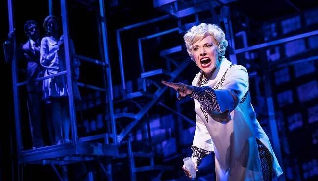 Emily Skinner singing “The Ladies Who Lunch”. She is wearing a short white wig and holding a champagne glass.