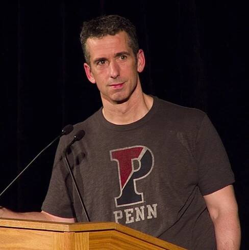 A 47-year-old white man is standing behing a lectern, looking to the camera's right; he has dark hair, and is wearing a grey t-shirt that says 