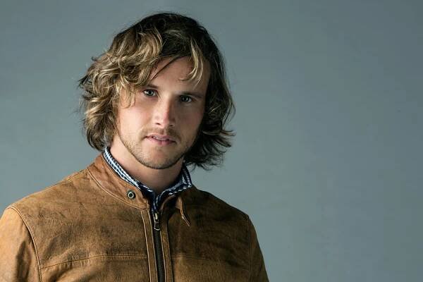 Young man with wavy hair wearing a brown suede jacket against a gray background.