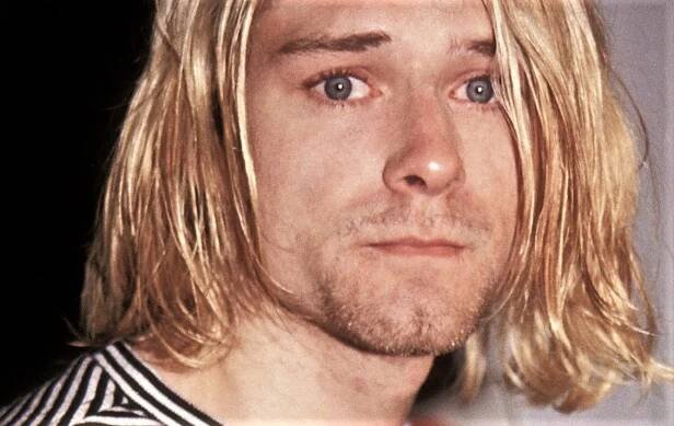 a man with long blonde hair and blue eyes looks at the camera while wearing a striped shirt