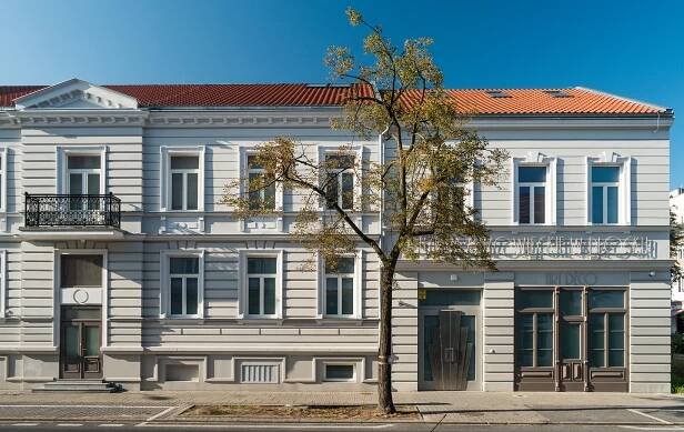 Budynek wystawy art deco Muzeum Mazowieckiego w Płocku przy ul. Kolegialnej 6, dwukondygnacyjna kamienica, stojąca w szeeregu innych, z czerwonym dachem i jasną fasadą Przed nią, na chodniku - drzewo.