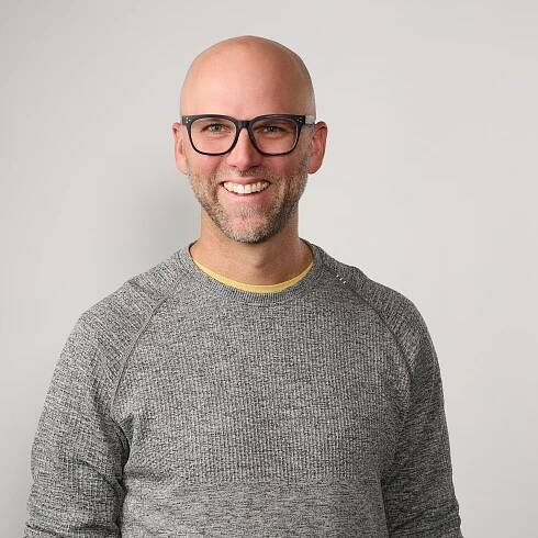 Casual headshot of a bald man with glasses, wearing a gray sweater, smiling warmly against a light gray background.