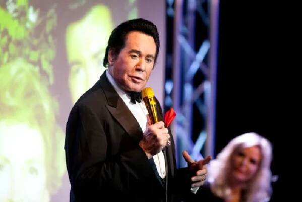 Wayne Newton performing on stage, dressed in a black tuxedo with a red pocket square, holding a golden microphone